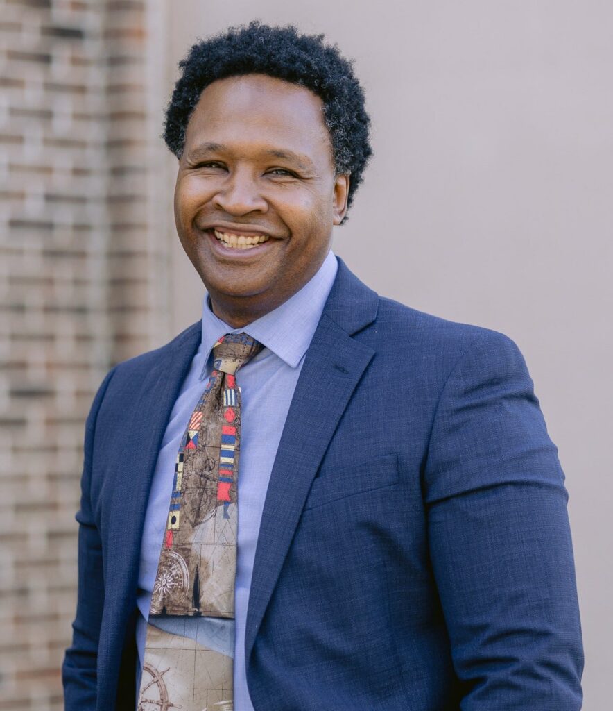Smiling man, Bryan Stringer, in a navy suit and patterned tie posing outdoors against a brick wall.